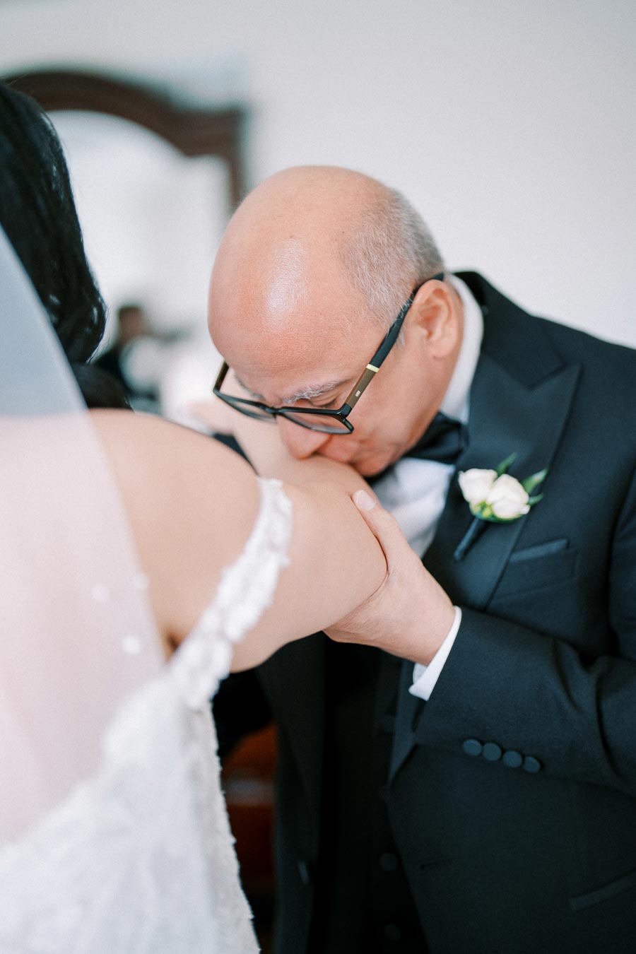 Elderly man in a suit tenderly kissing the arm of a bride in a white wedding dress, capturing a sentimental moment during a wedding ceremony.
