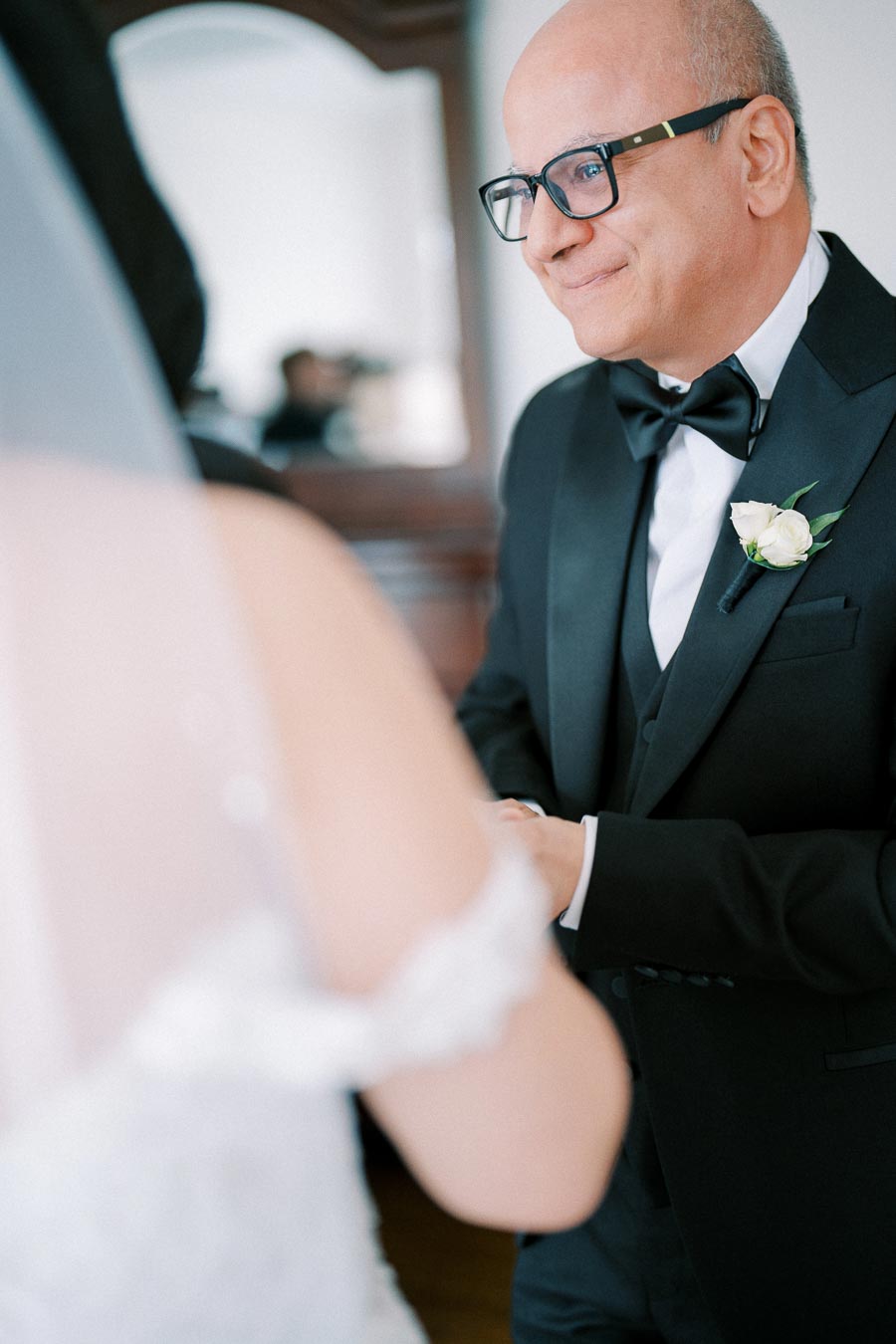 A groom in a black tuxedo and glasses smiling as he holds hands with a bride, who is wearing a white dress, during a wedding ceremony.