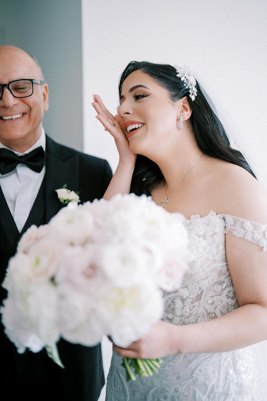 Joyful bride in a lace wedding dress, holding a bouquet of white flowers, shares a happy moment with a smiling man in a tuxedo, capturing the essence of a wedding day celebration.