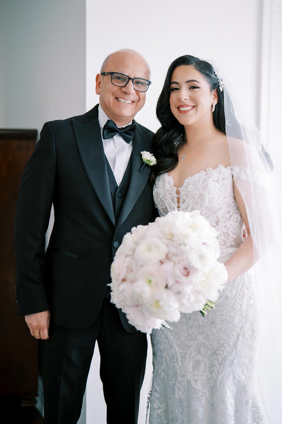 Father and bride smiling on wedding day, wearing elegant attire; bride holds a bouquet of white flowers.