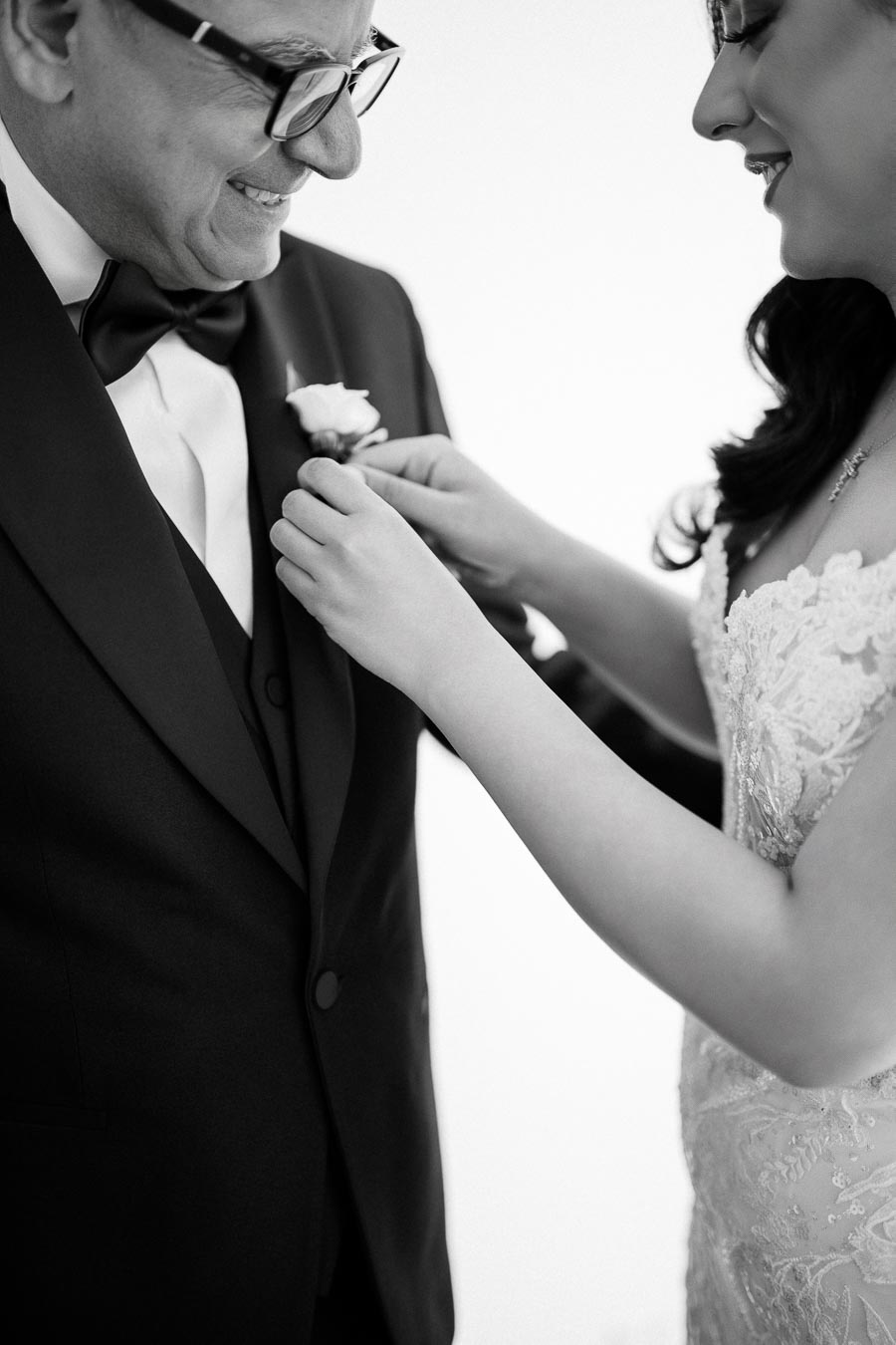 A bride in a lace gown pins a boutonnière on a smiling man in a tuxedo, highlighting an intimate wedding preparation moment.
