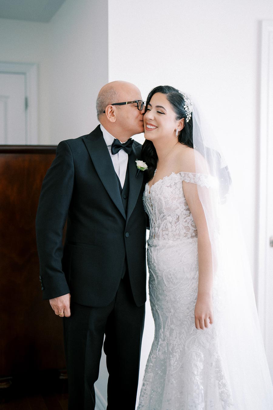 Father affectionately kissing bride on the cheek, both smiling, in elegant wedding attire.