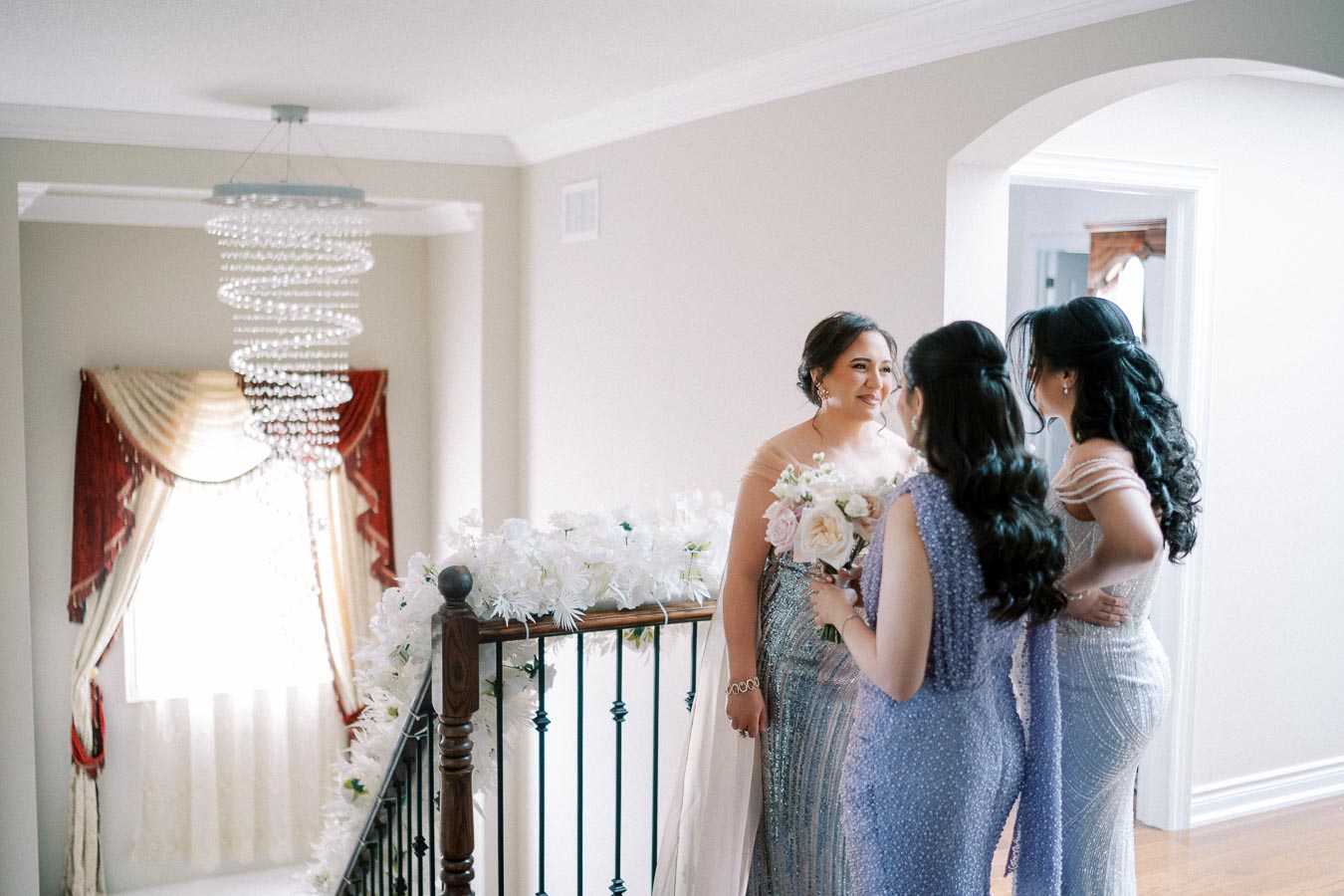 Elegant wedding preparation scene with a bride and bridesmaids in shimmering dresses, holding a bouquet, standing on a decorated staircase with a cascading chandelier in background.