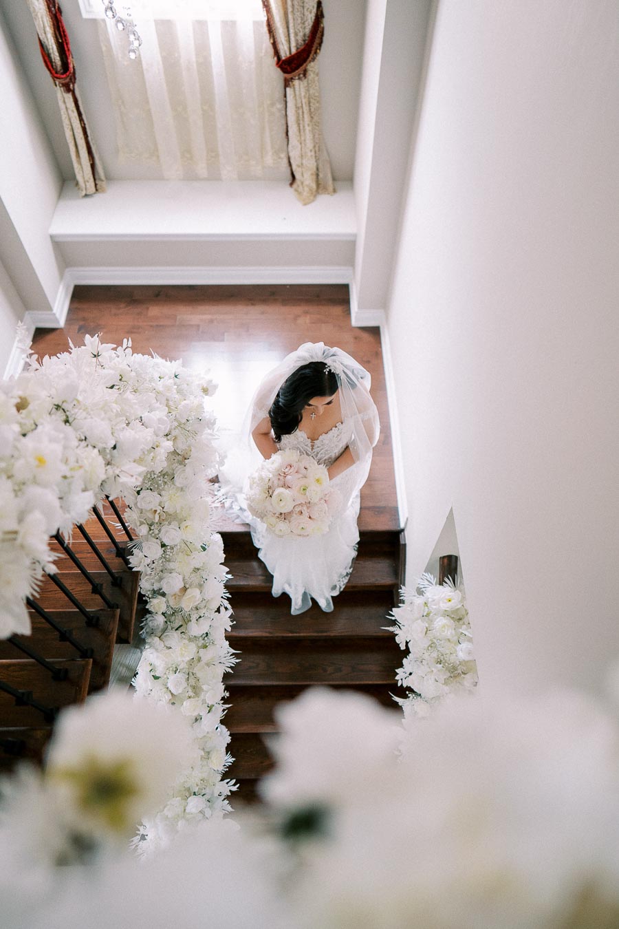 A bride in a white wedding dress and veil holds a bouquet of light-colored flowers while descending a staircase decorated with cascading white floral arrangements.