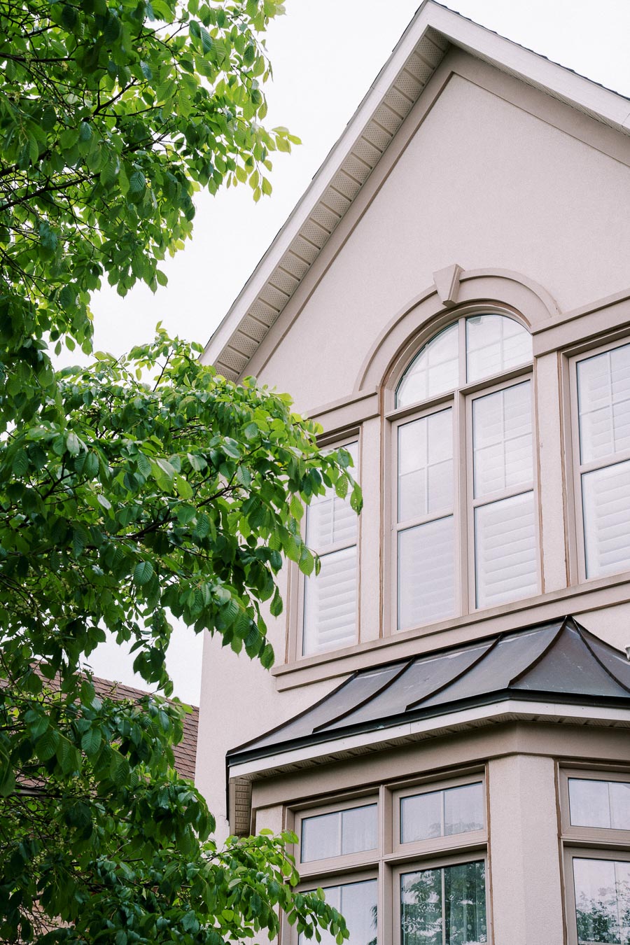 Gable roof of a modern house with large windows and shutters, surrounded by leafy green trees.