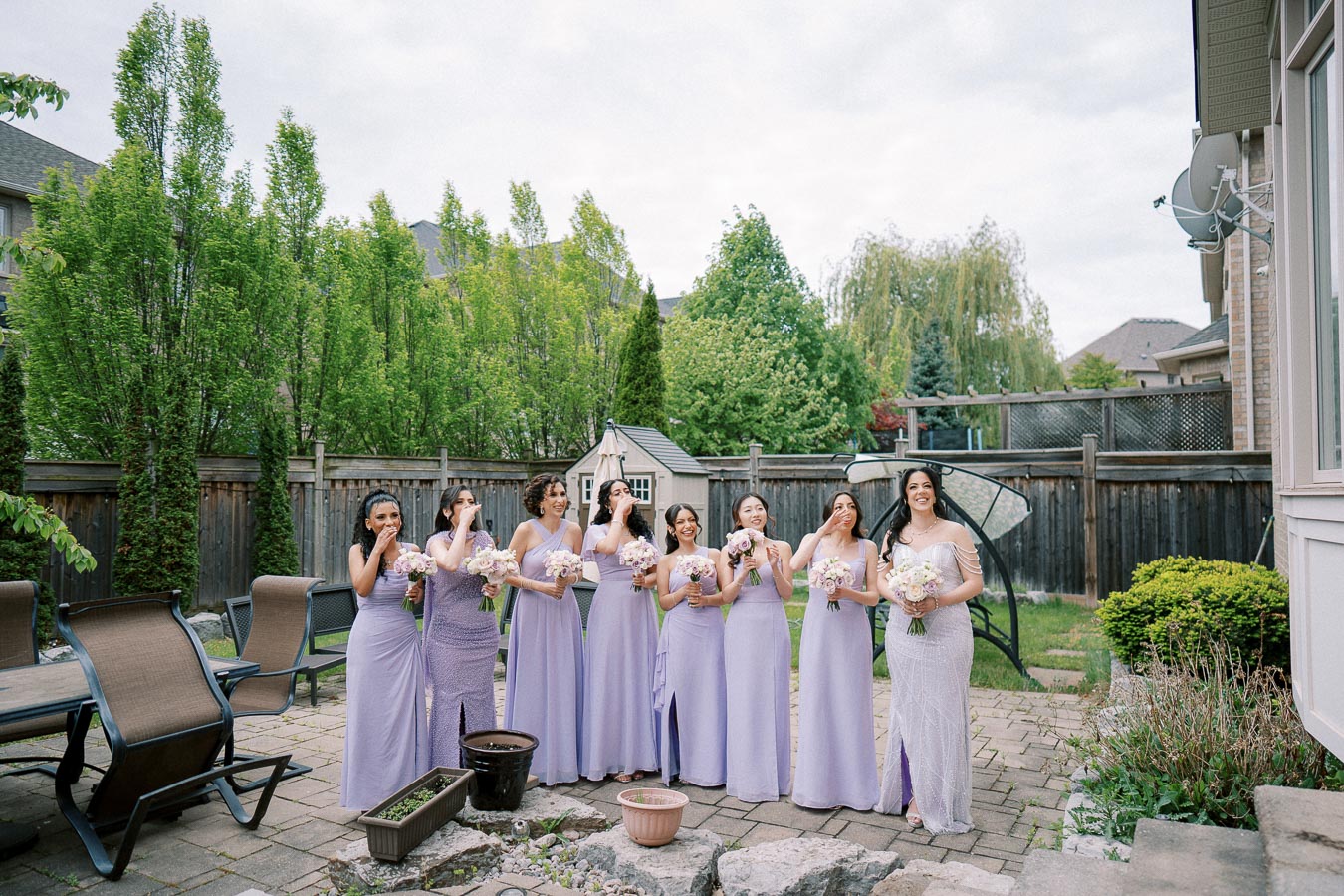 Bridesmaids in lavender dresses standing in a garden, smiling and holding bouquets, with lush greenery and patio furniture in the background.
