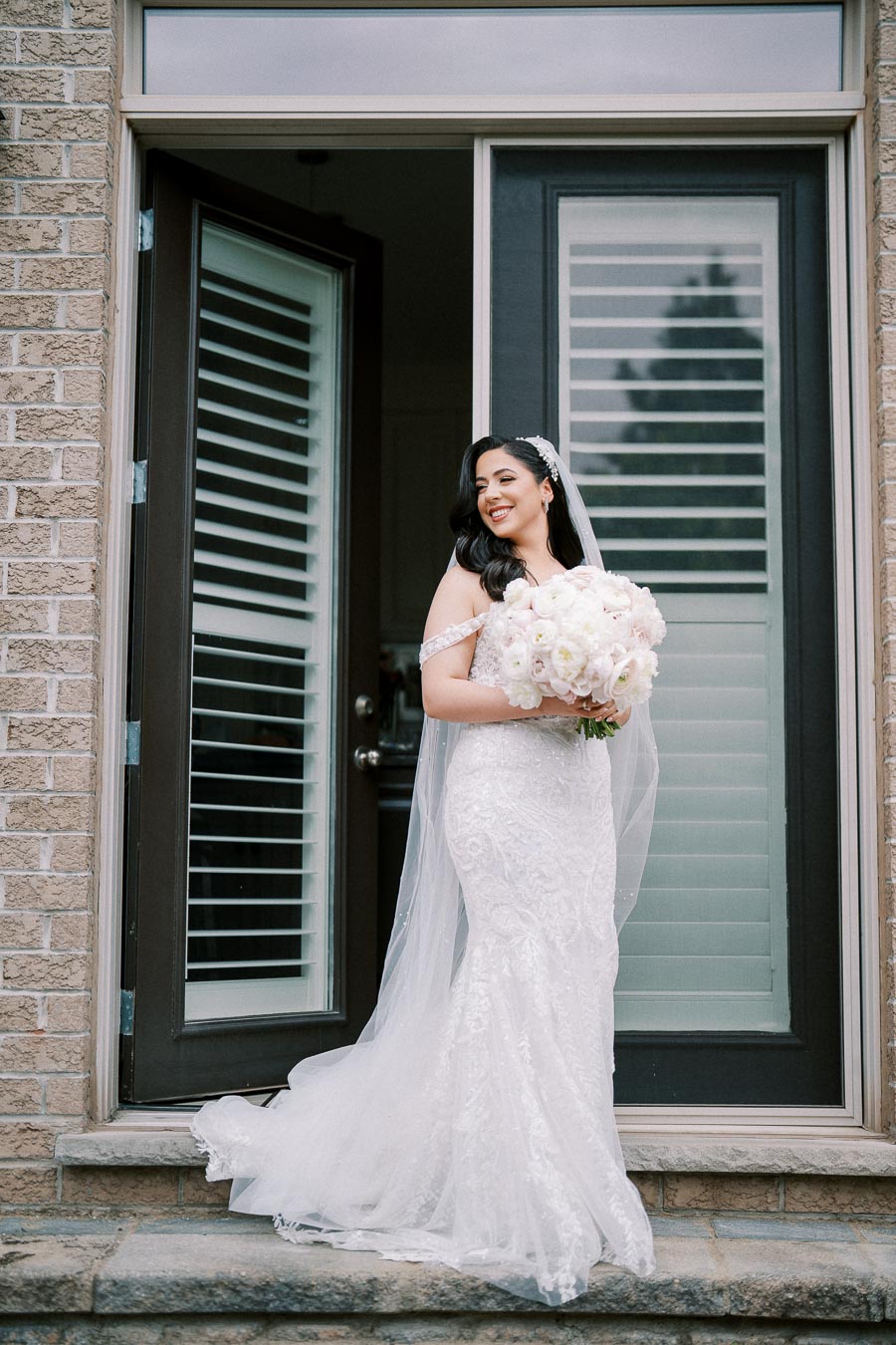 Smiling bride in elegant white lace wedding dress, holding a bouquet of white and blush flowers, standing in a doorframe.