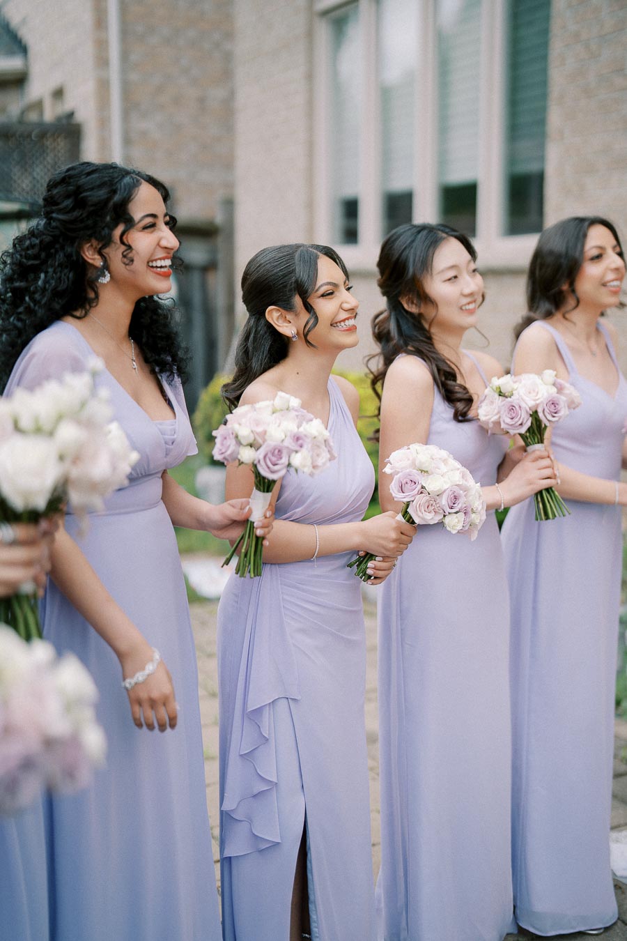 Four bridesmaids in matching lavender dresses smile while holding bouquets of pink and white flowers, standing outside a building.