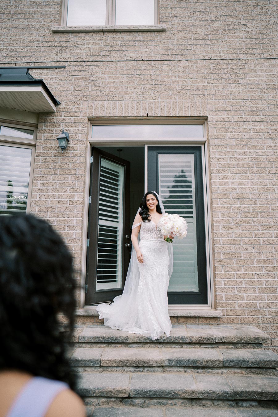 A bride in an elegant white lace wedding dress holding a bouquet of white flowers, smiling as she steps out of a house with brick exterior on a special occasion.