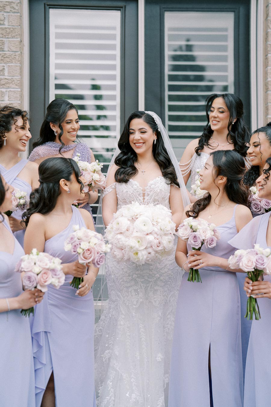 Bridal party in lavender dresses surrounding the bride in a white lace gown holding a bouquet of roses, posing happily in front of a modern building with large windows.