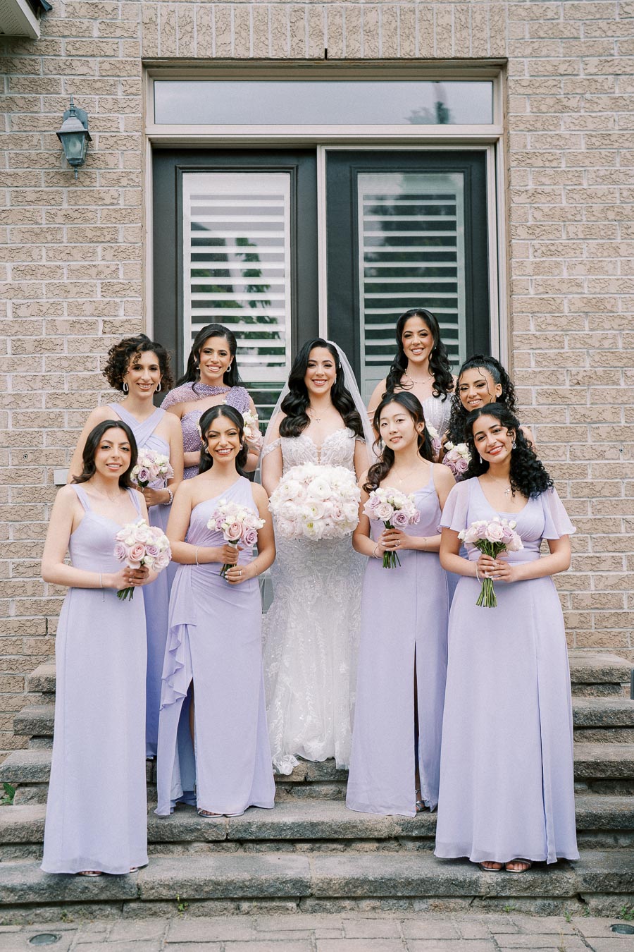 A bride surrounded by eight bridesmaids in matching lavender dresses, all holding bouquets, standing on stone steps in front of a brick building.