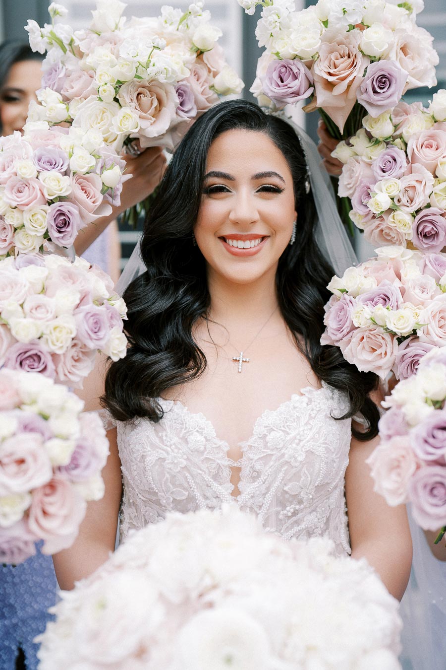 Smiling bride surrounded by pastel-colored rose bouquets, wearing a lace wedding dress and veil, with a silver cross necklace.