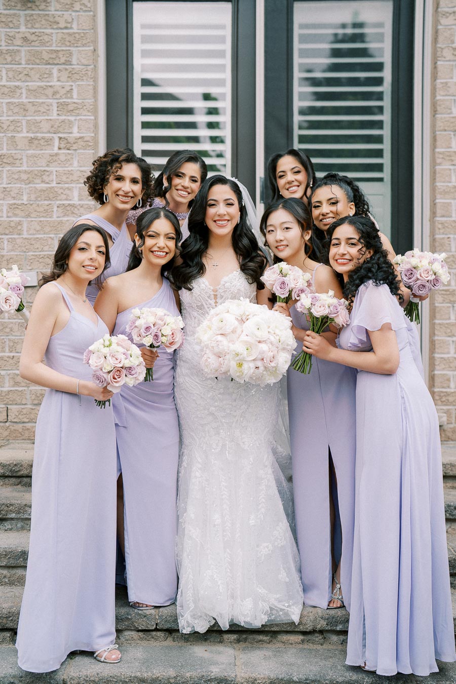 Bridal party photo featuring a smiling bride in a white lace gown and eight bridesmaids in lavender dresses, all holding bouquets of pastel roses, posed in front of a brick building with large windows.