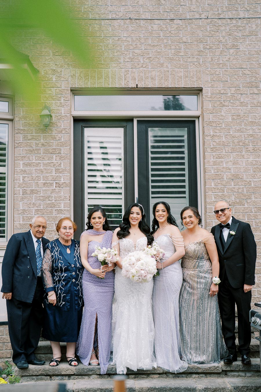A family group portrait at a wedding, with the bride in a white gown holding a bouquet, surrounded by elegantly dressed family members, posing in front of a house with brick walls and large windows.