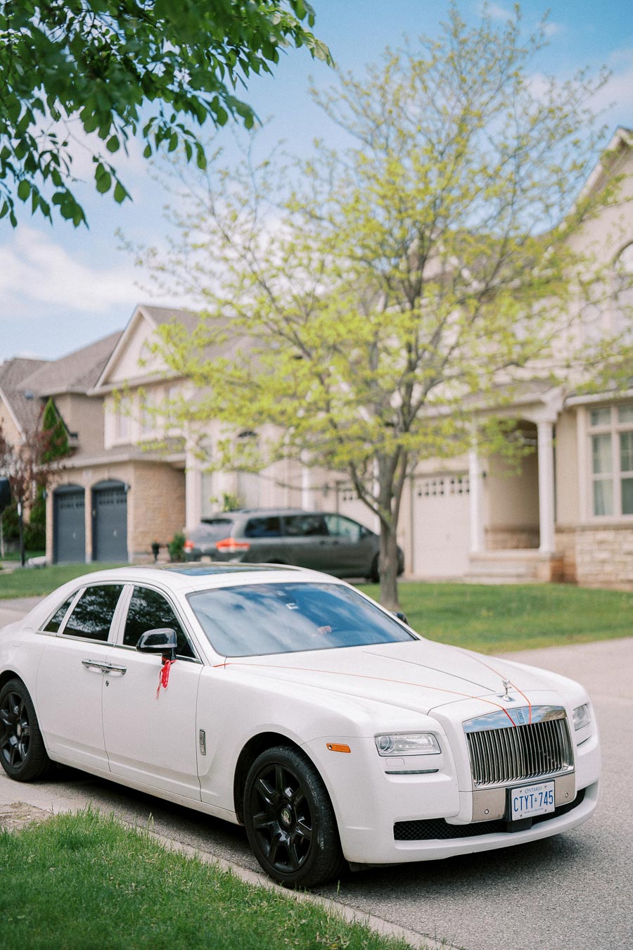 A luxurious white car with black rims parked in a suburban neighborhood, surrounded by green trees and elegant houses, under a clear blue sky.