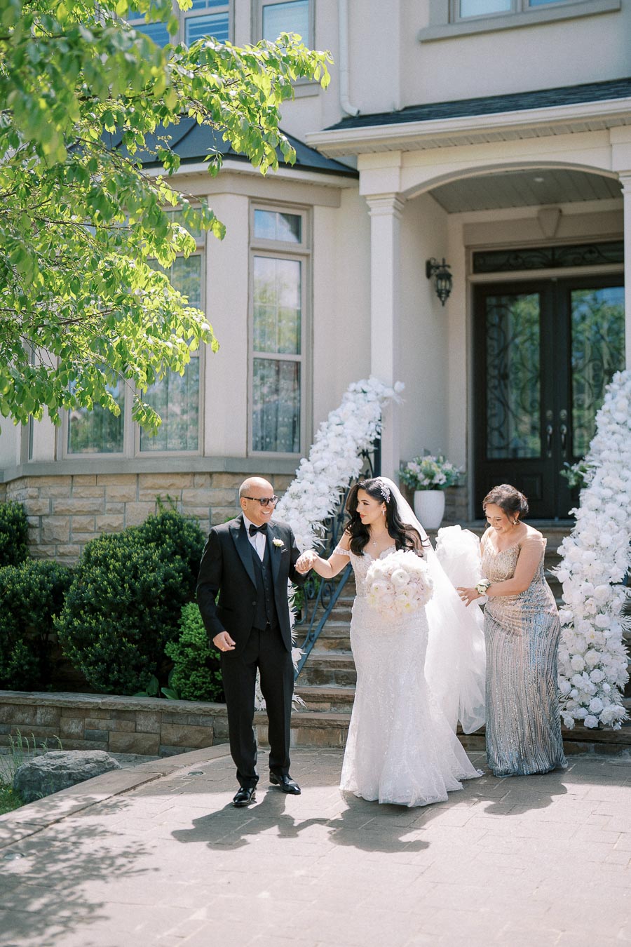 Bride in a white lace gown accompanied by two elegantly dressed individuals, standing in front of a beautifully decorated entrance with white flowers, surrounded by greenery on a sunny day.