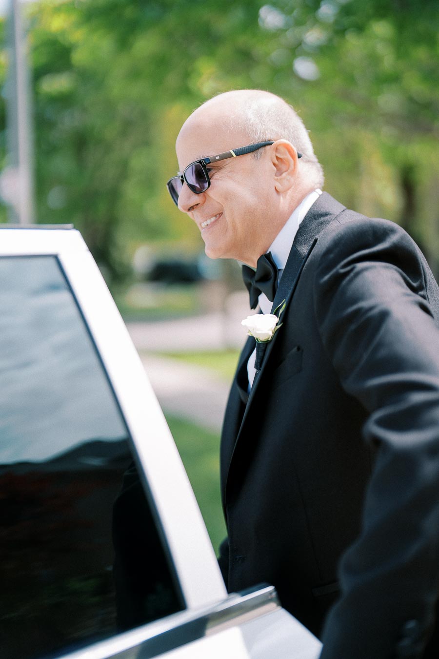 A smiling man in a tuxedo and sunglasses getting into a car on a sunny day, with trees in the background.