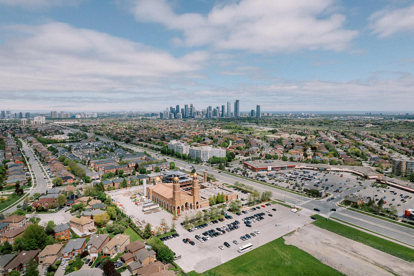 Aerial view of a suburban neighborhood with a prominent church in the foreground, parking lots, residential streets with houses, and a distant urban skyline under a partly cloudy sky.
