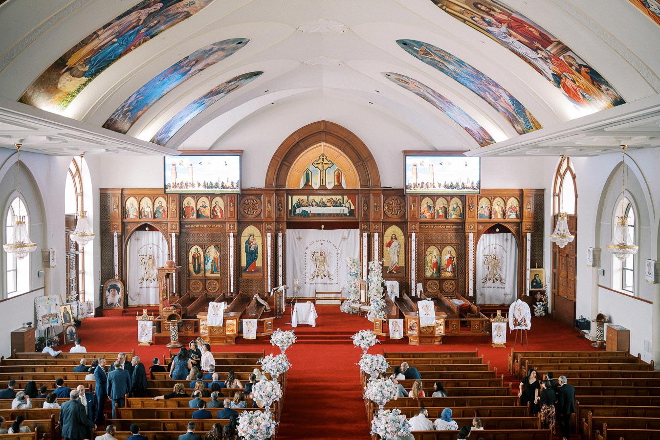 Interior of a beautifully decorated church featuring ornate wooden carvings and colorful religious artwork. The central aisle is adorned with floral arrangements, leading to an intricately designed altar. Attendees are seated on wooden pews, absorbing the serene and majestic atmosphere of the sacred space.