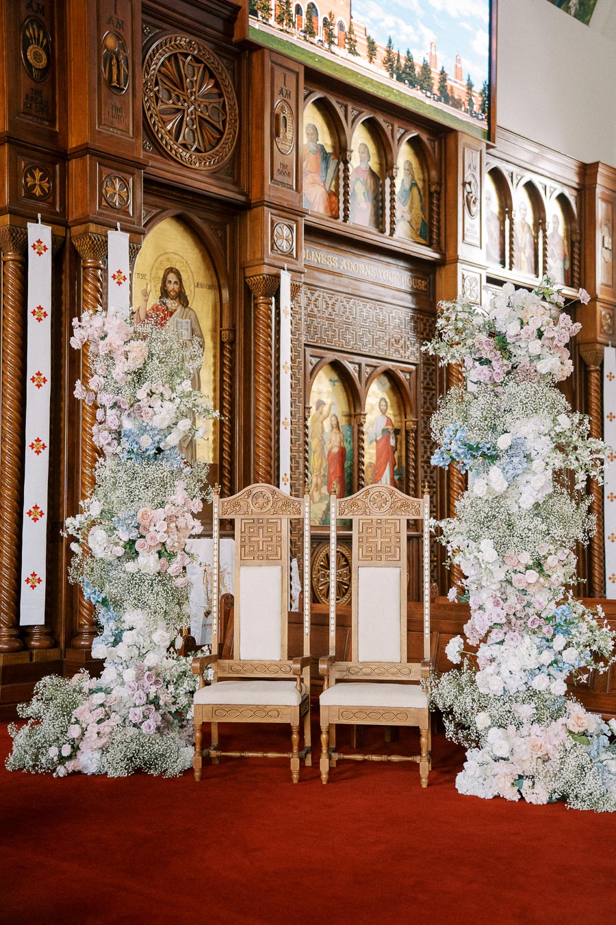 Two intricately carved wooden chairs sit in front of a beautifully adorned altar in a church, surrounded by elegant floral arrangements featuring soft pink and white blooms.