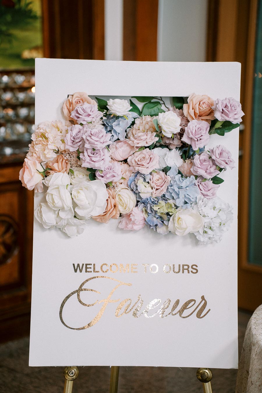 Wedding welcome sign decorated with pastel-colored flowers, featuring the message 'Welcome to Our Forever' in elegant gold lettering.