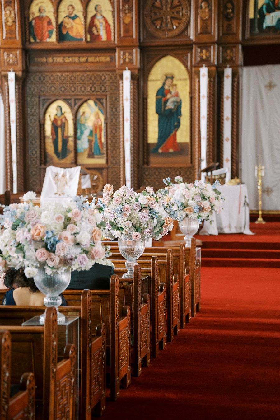 Ornate church interior with wooden pews adorned with elegant floral arrangements, featuring pastel roses and hydrangeas; intricate religious art and iconography in the background.
