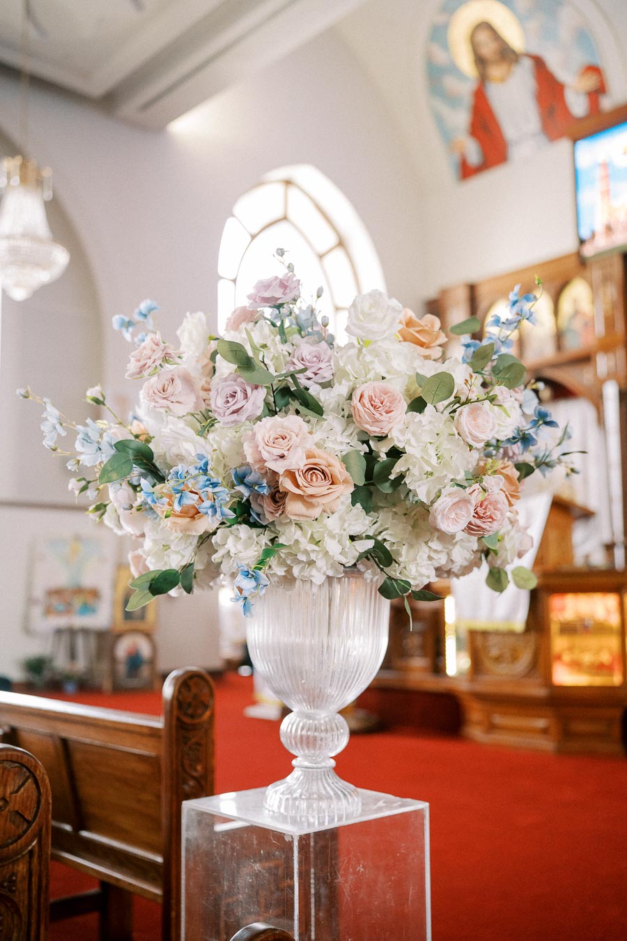 Elegant floral arrangement featuring pink and white roses with blue accents in a glass vase inside a church with red carpeting and stained glass windows.