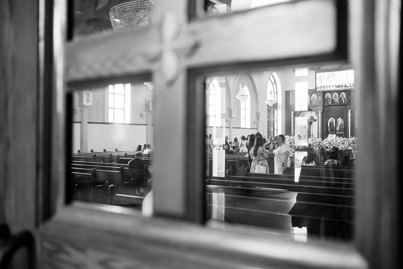 Black and white photo of a church interior, viewed through a door window, showing people seated in pews and standing at the altar area.