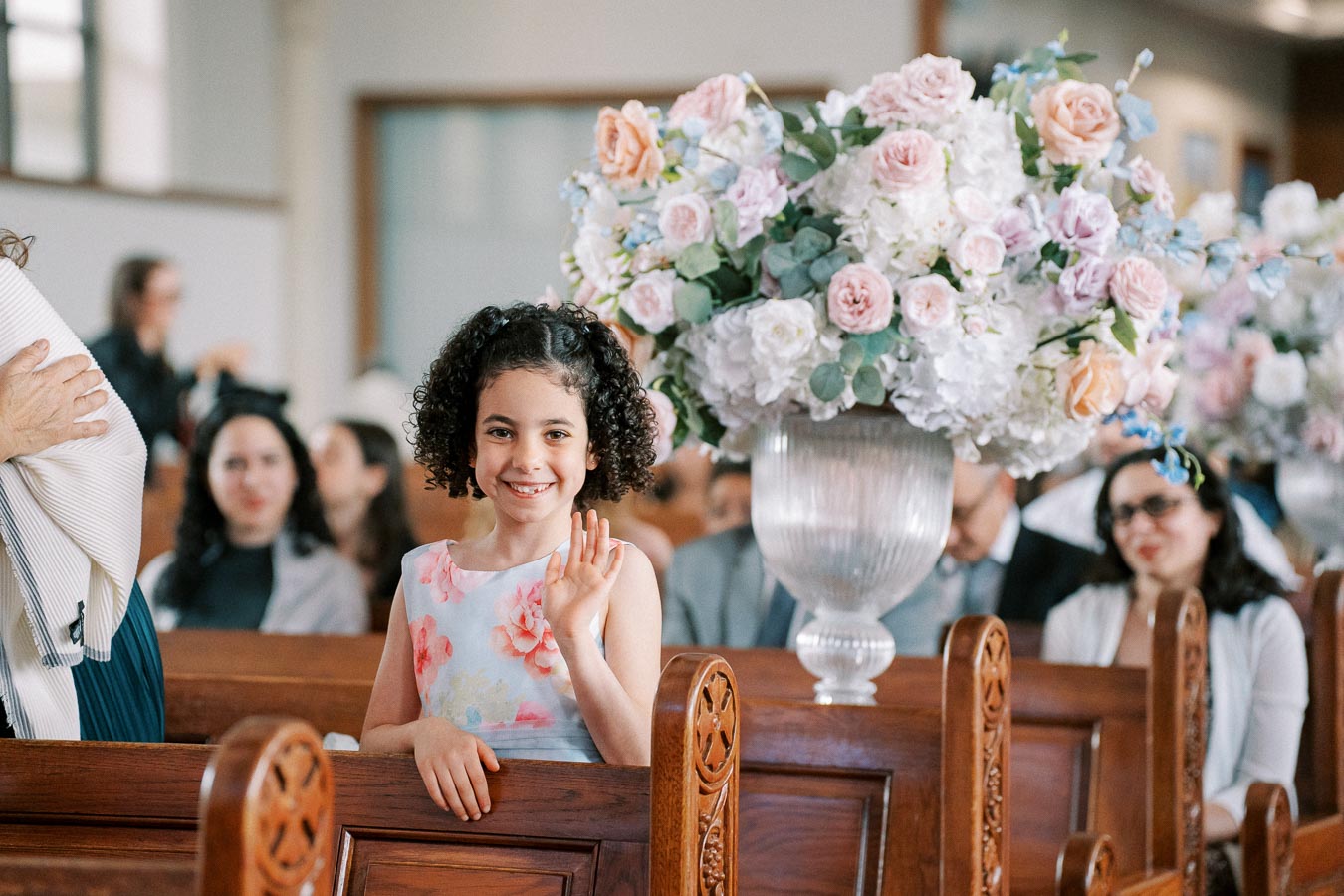 A young girl with curly hair in a floral dress smiling and waving at a formal event, seated in a wooden pew with a large bouquet of pink and white flowers in the background.