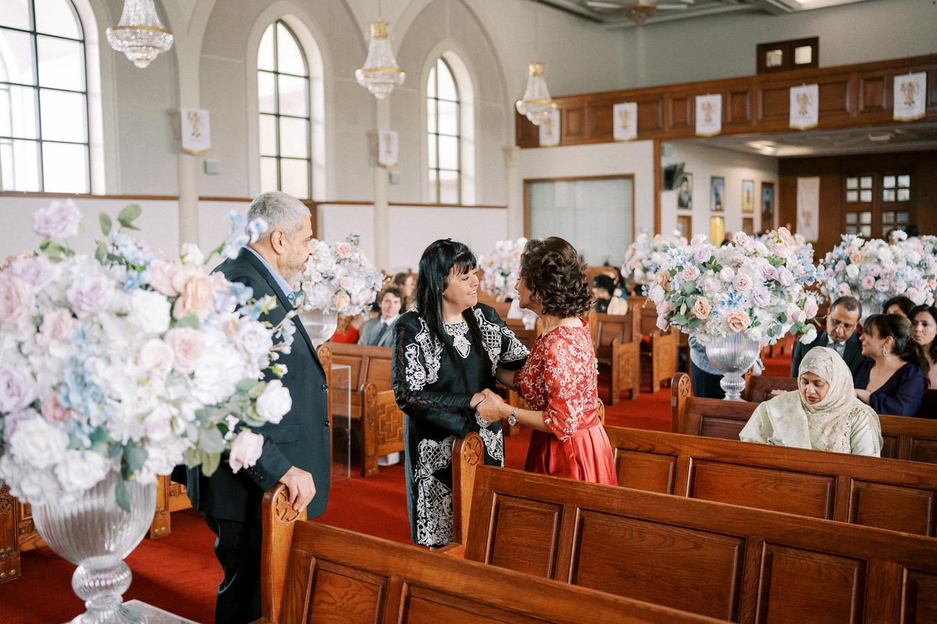 A couple greeting each other in a decorated church interior, with large floral arrangements and wooden pews in the background.