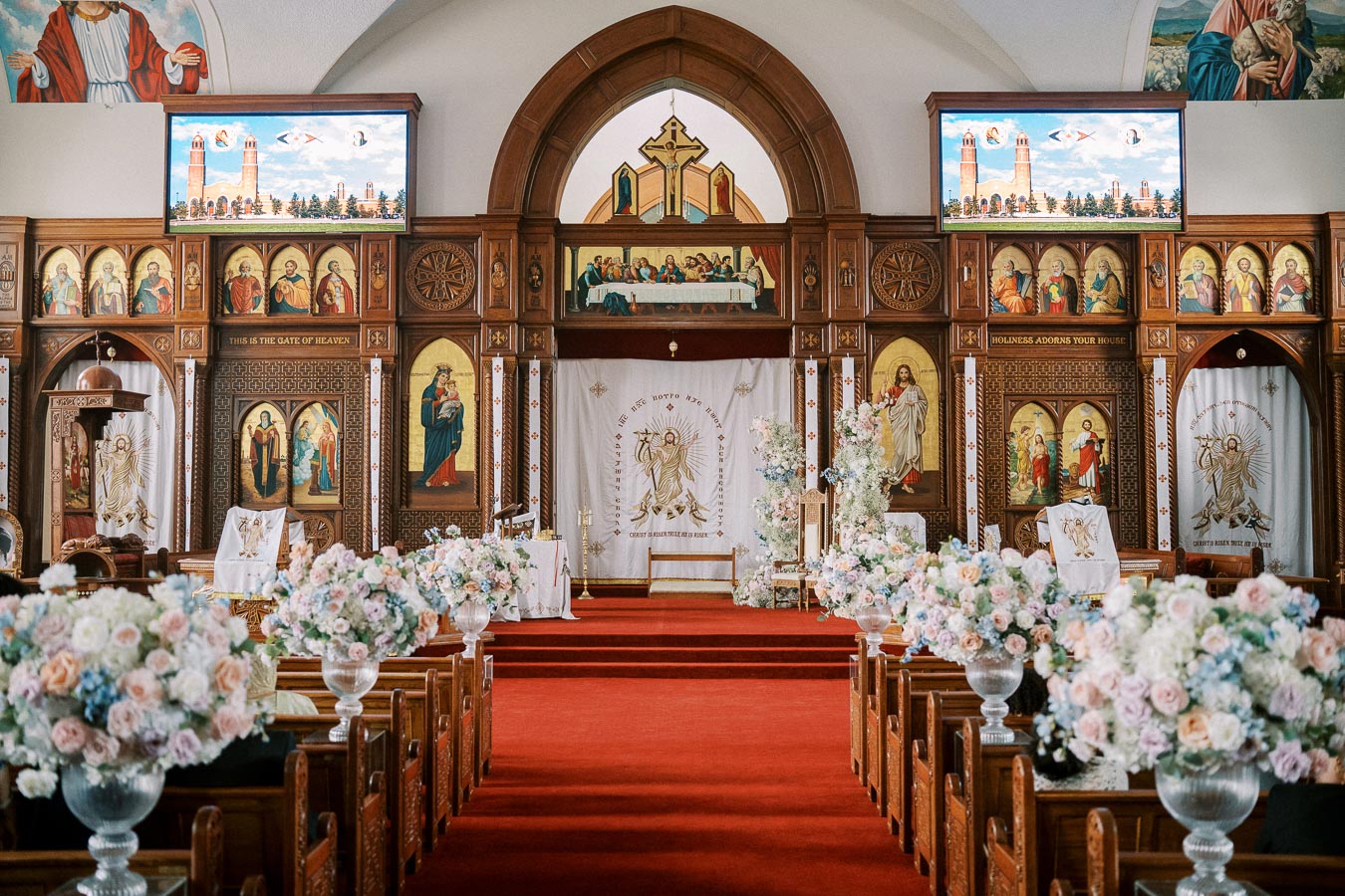 Interior of an ornate church featuring a richly decorated altar with religious icons and artwork, surrounded by pews adorned with floral arrangements, beneath high ceilings with intricate detailing.