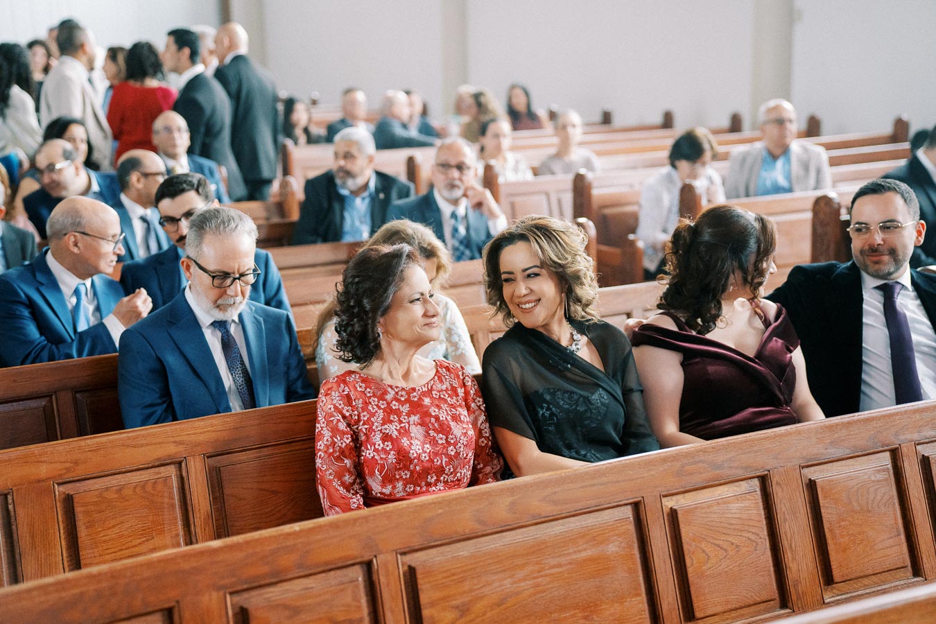 A group of well-dressed people sitting in wooden pews, socializing before a formal event in a church setting.
