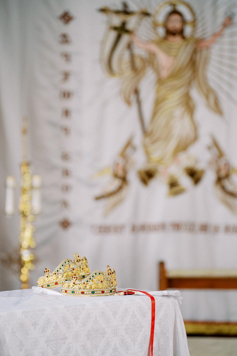 Golden crowns adorned with colorful jewels on a white altar cloth beneath a religious tapestry featuring angelic figures.