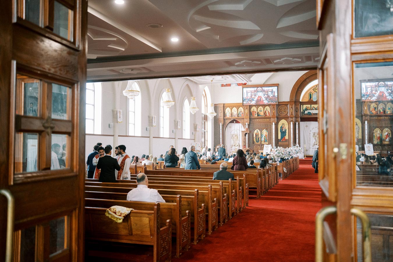 Interior of a church during a service, featuring attendees seated on wooden pews facing an ornate altar with religious art and icons, under soft chandelier lighting.