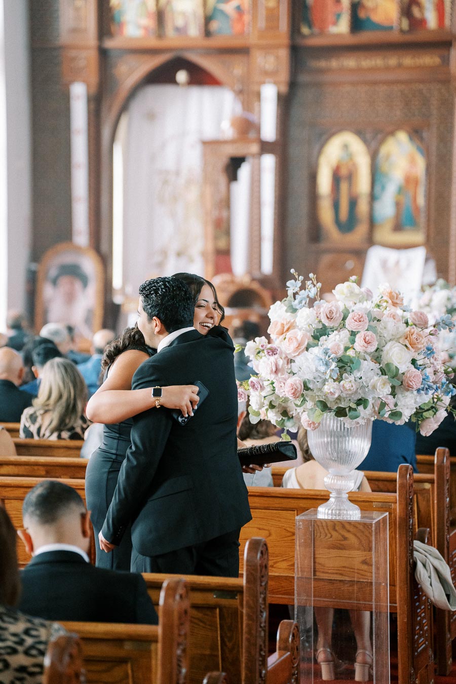 Two people embracing inside a church decorated with large floral arrangements, surrounded by seated guests during a ceremony.
