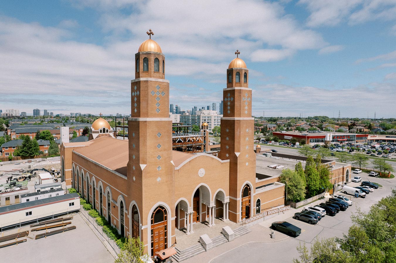 Aerial view of a large, modern church with two tall bell towers and a gold dome, set against an urban backdrop with residential homes and commercial buildings under a partly cloudy sky.