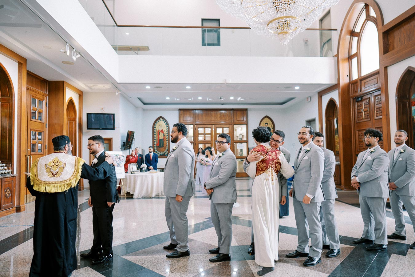 A group of men in suits standing in line inside an elegant venue, with one man hugging a person in ceremonial robes. The atmosphere is formal and ceremonial, featuring wooden paneling, high ceilings, and decorative elements.