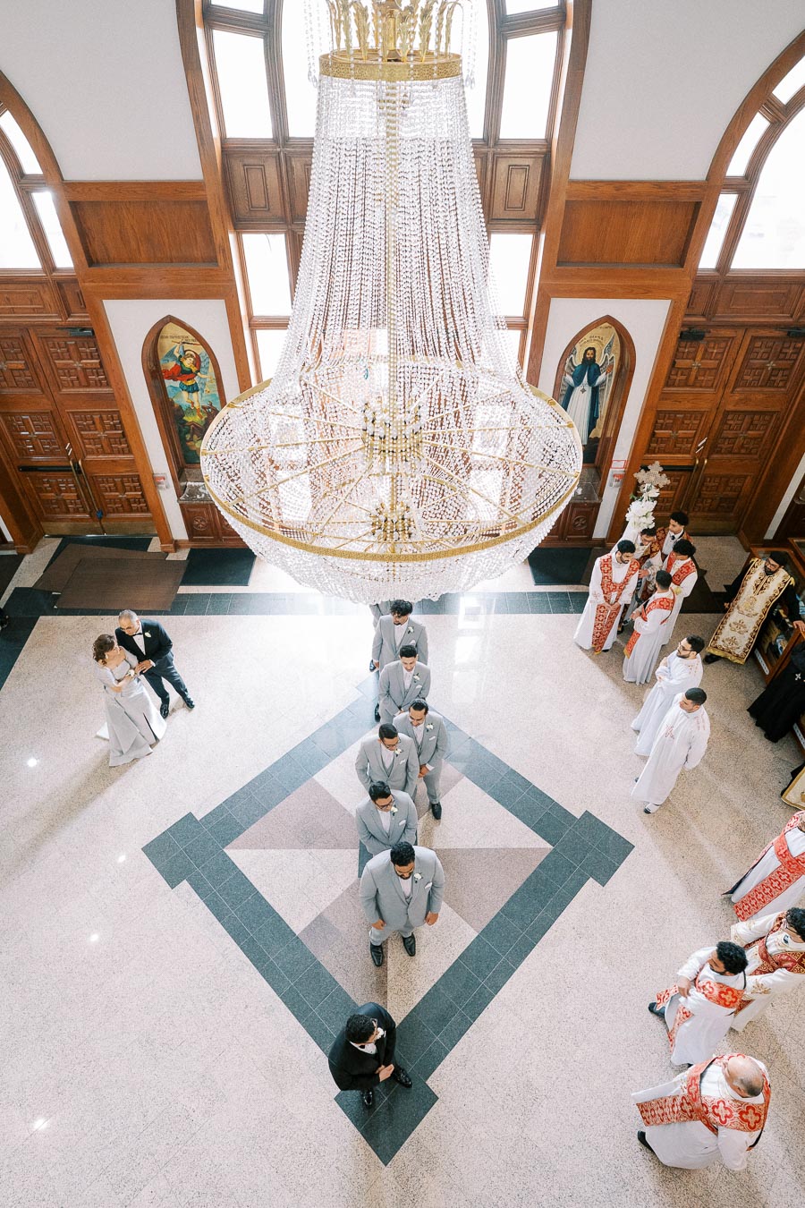 Aerial view of a church interior with a large crystal chandelier and people gathered for a wedding ceremony. Groomsmen in grey suits are lined up, and priests in ceremonial robes are visible. The setting features ornate woodwork and religious artwork.
