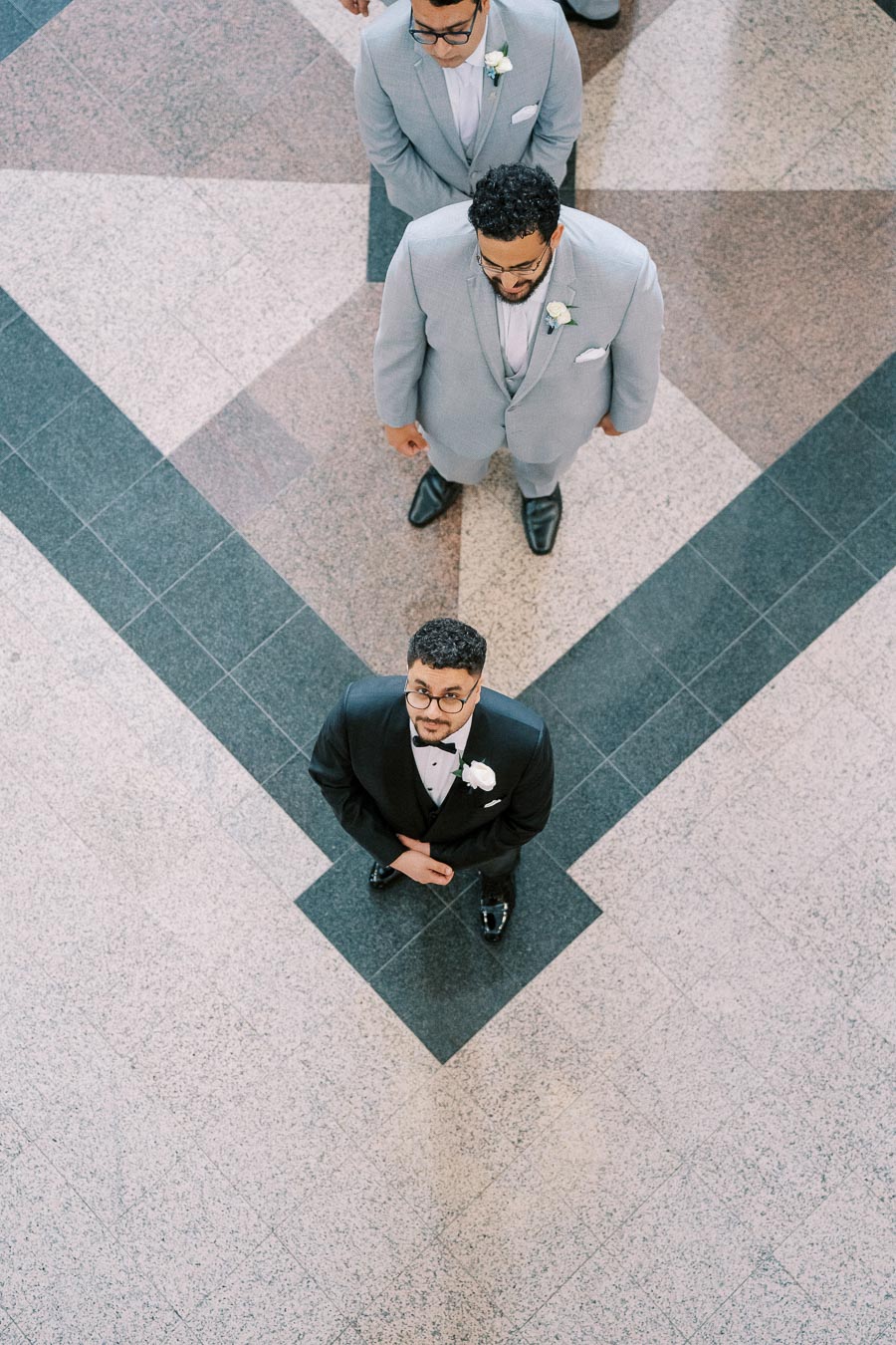 Overhead view of three men in formal attire standing in a geometric-patterned lobby; the groom wears a black tuxedo with a white boutonniere, while the two groomsmen are in matching light gray suits.