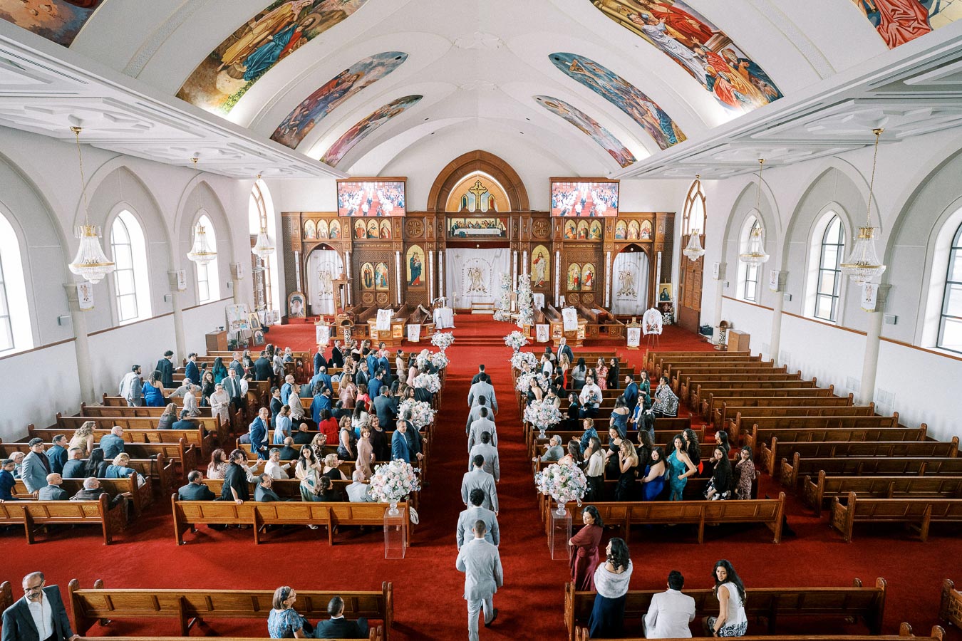 A beautifully decorated church interior filled with guests attending a wedding ceremony. The image shows a red carpet aisle, floral arrangements, and detailed ceiling artwork, creating a serene and joyous atmosphere.