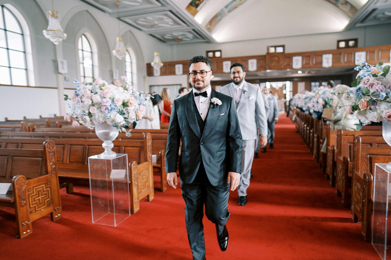 A groom and groomsmen walking down the aisle in a beautifully decorated church with floral arrangements and wooden pews.
