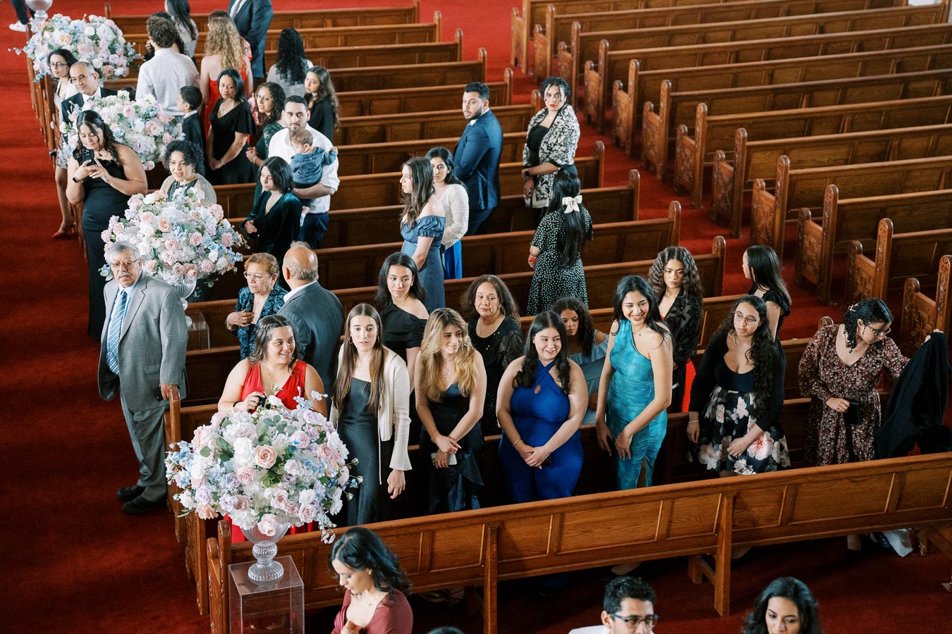 A diverse group of people dressed in formal attire stand in the pews of a beautifully decorated church, with elegant floral arrangements lining the aisle, suggesting a wedding or special ceremony.