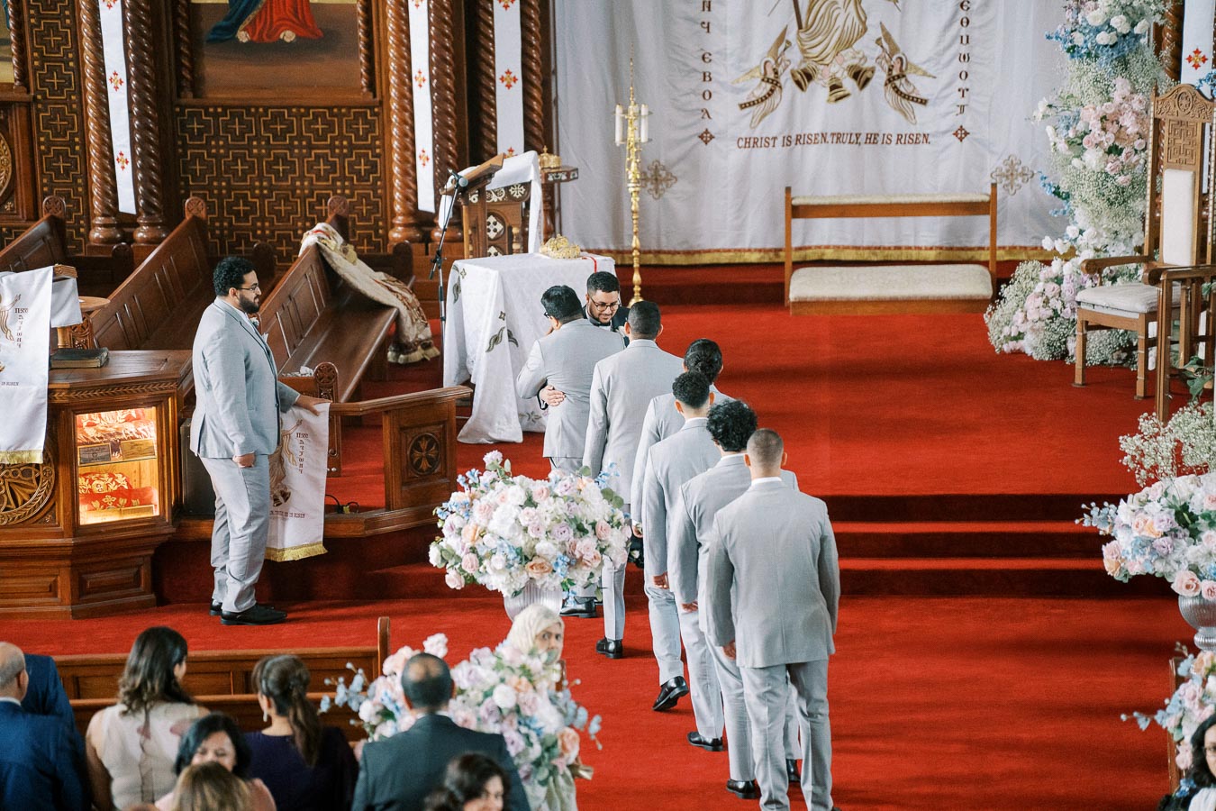 A group of groomsmen in light gray suits stands in line at the front of an ornately decorated church during a wedding ceremony, with floral arrangements and an altar adorned with religious symbols.