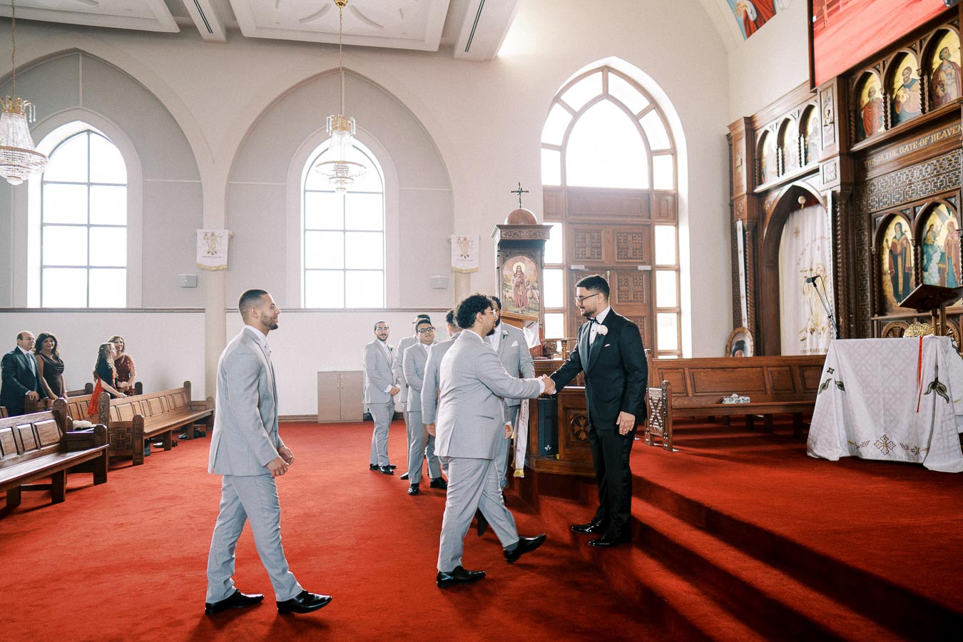 Groomsmen in light gray suits line up to greet a man in a black suit and bow tie, exchanging a handshake in a beautifully decorated church with wooden pews, red carpet, and arched windows.