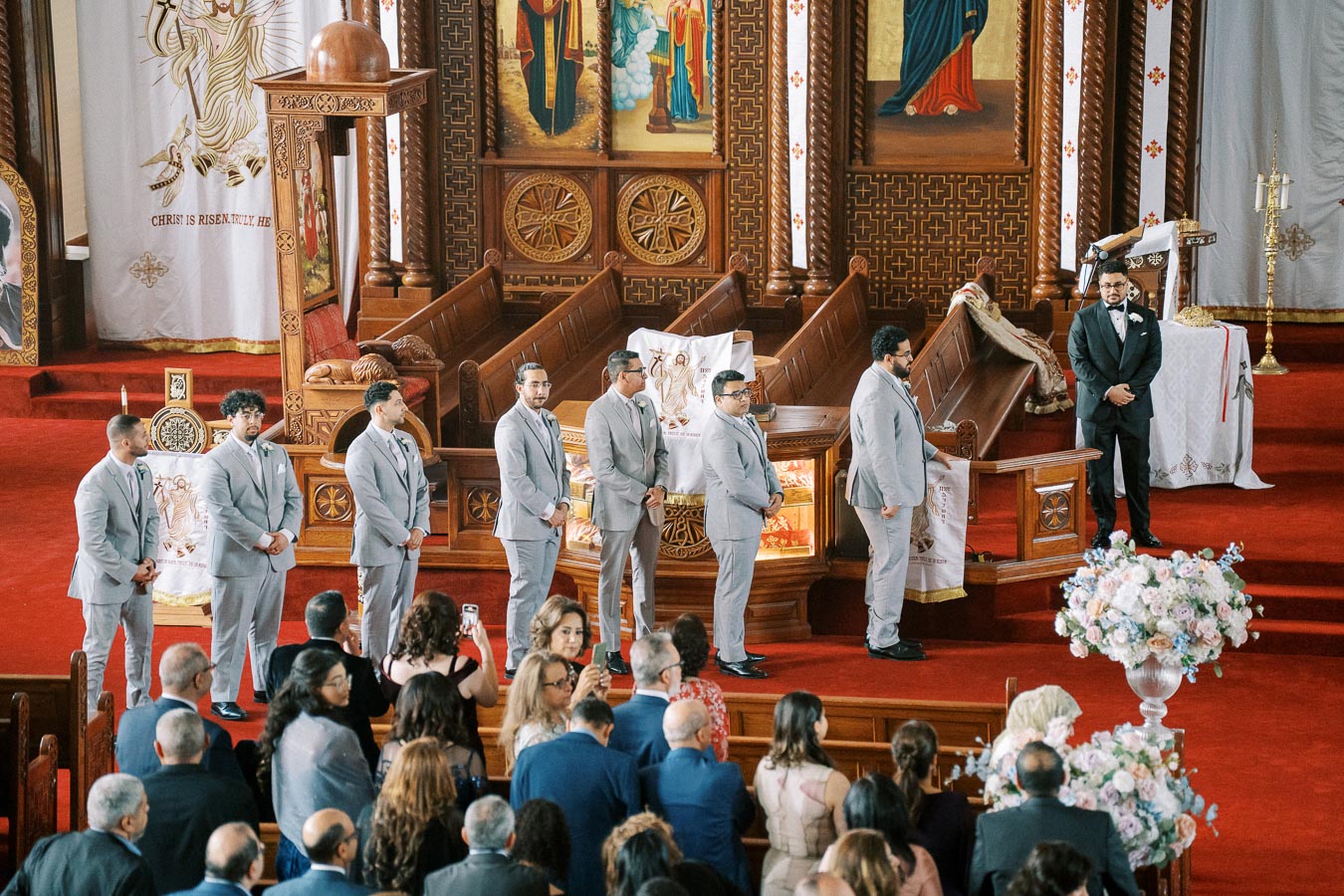 Groomsmen in matching gray suits stand in a row inside an ornately decorated church, featuring detailed wooden structures and religious imagery, as a crowd of guests watches the wedding ceremony.