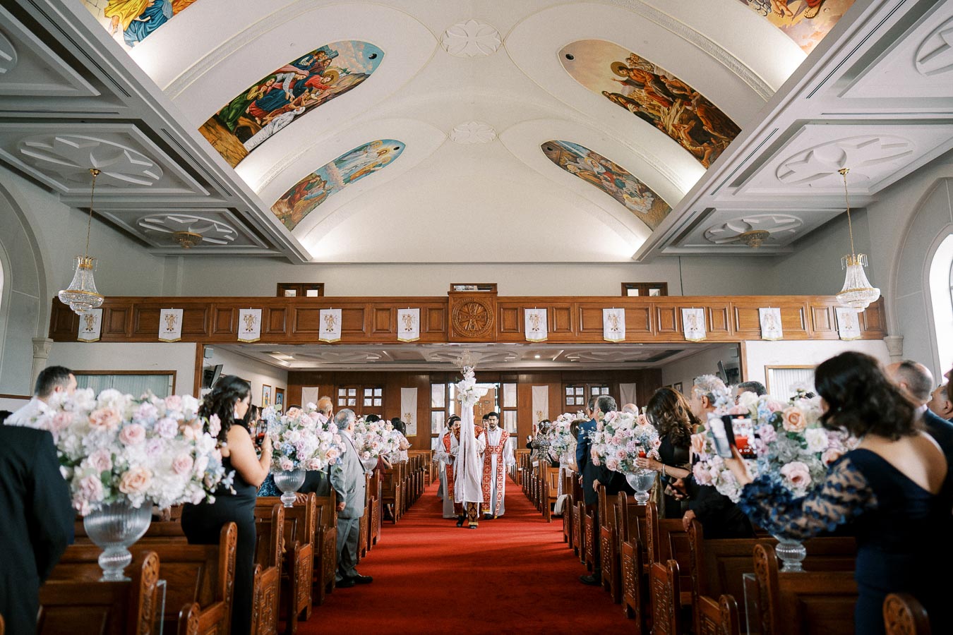 A beautifully decorated church interior during a wedding ceremony, with guests seated along the aisle holding floral arrangements and priests approaching the altar under a ceiling adorned with religious artwork.