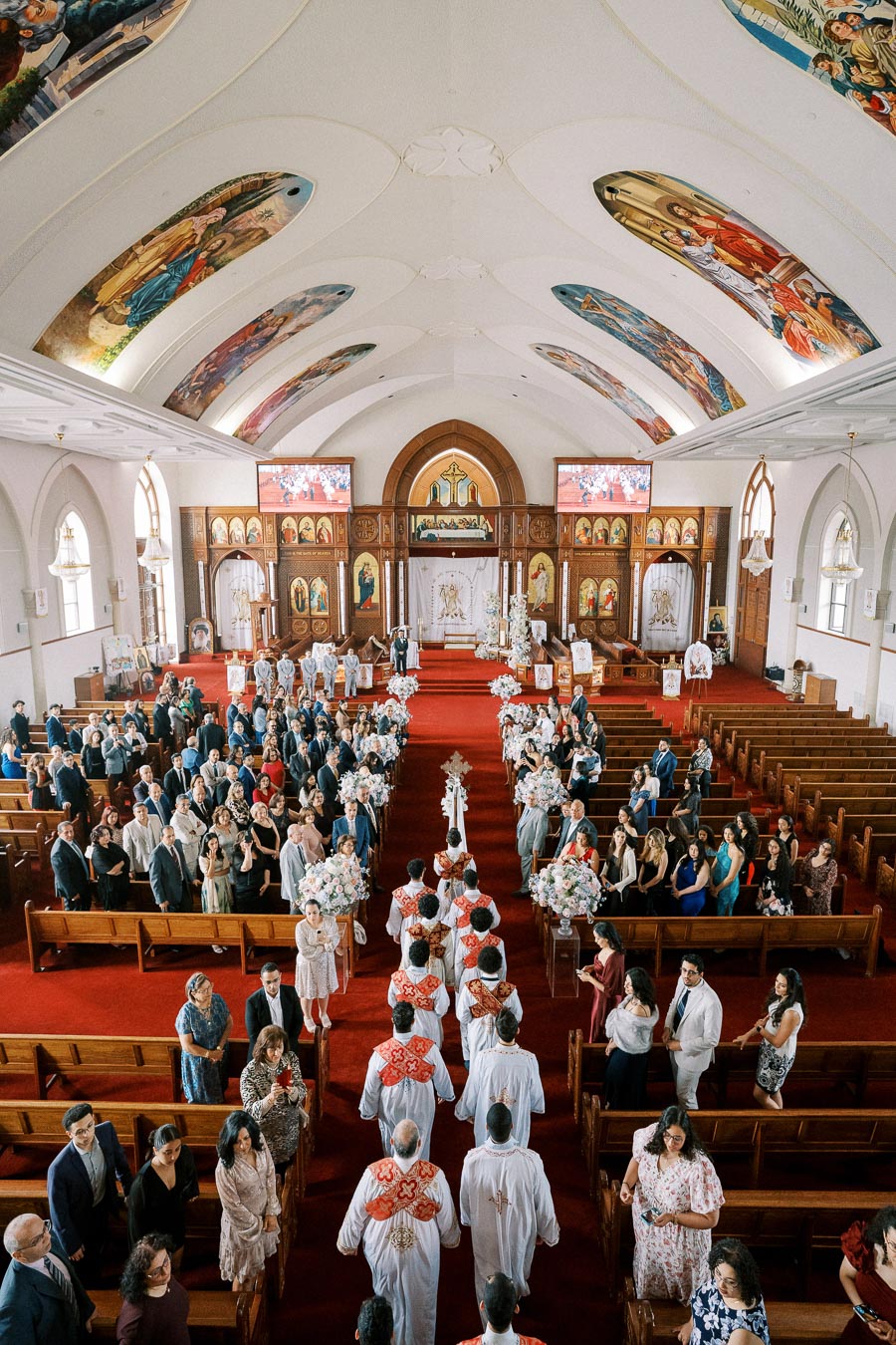 Alt text: Interior of a church during a wedding ceremony with a procession of clergy in white robes and red sashes walking down the aisle, surrounded by seated guests and elaborately decorated with floral arrangements and religious iconography.