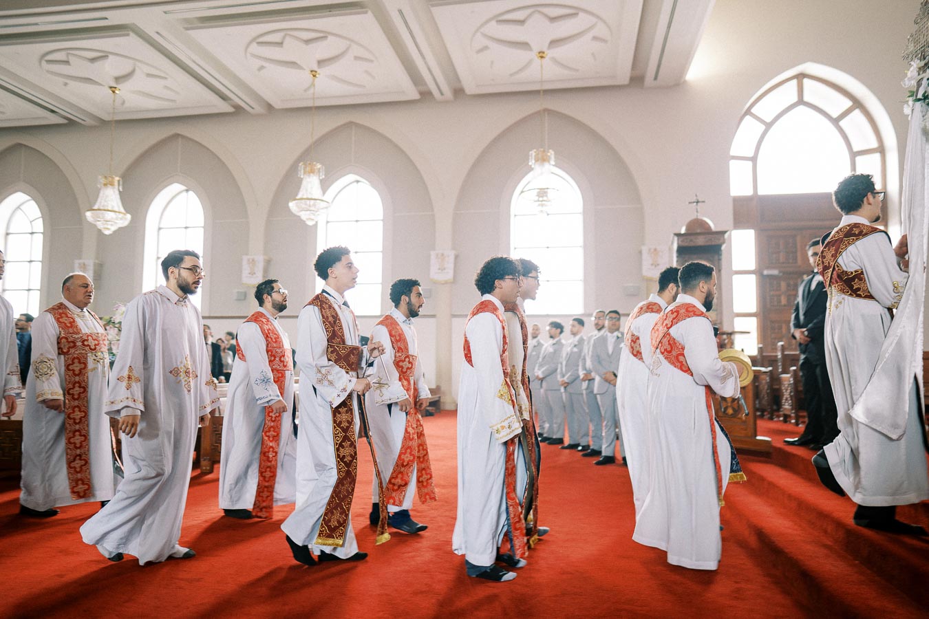 Priests in white and red liturgical garments during a procession inside a church with arched windows and chandeliers.