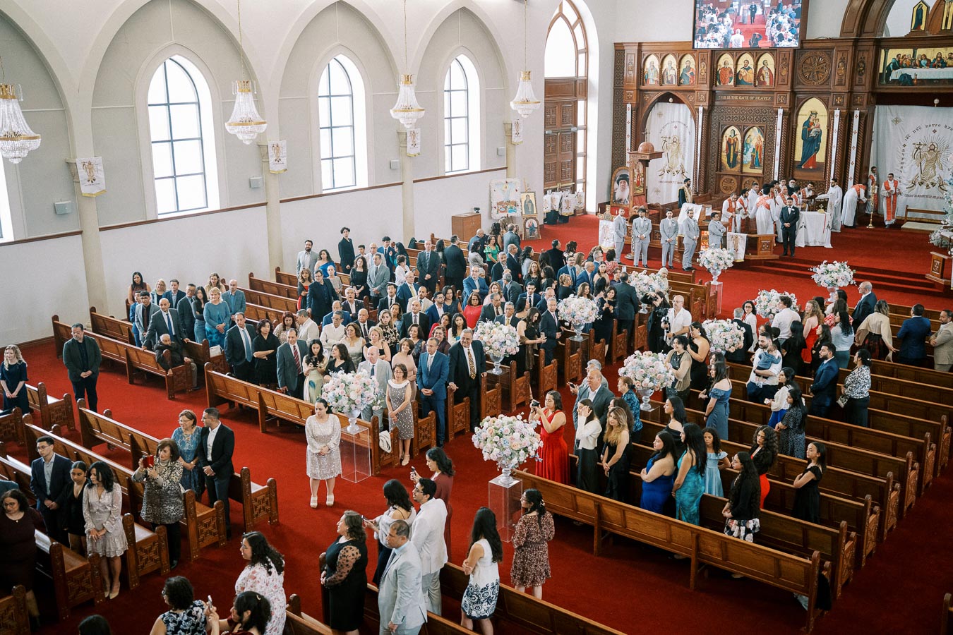 A large congregation gathered inside a beautifully decorated church with arched windows, elegant chandeliers, and floral arrangements; people are formally dressed, with the ceremony occurring at the altar in front.