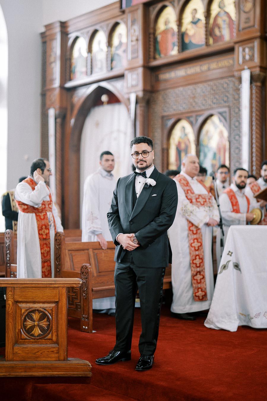 A man in a black tuxedo stands in a church with ornate wooden décor and religious figures, surrounded by clergy wearing white and red robes.